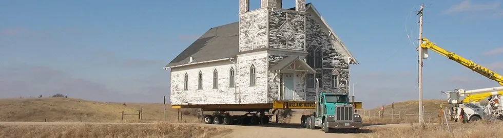 White wooden church with a bell tower on a trailer behind a teal truck