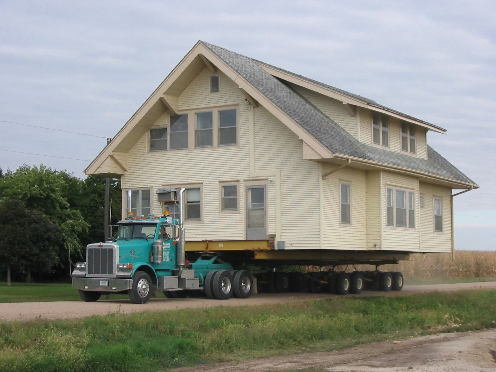 Cream craftsman-style house on a trailer behind a teal Peterbilt on a gravel road