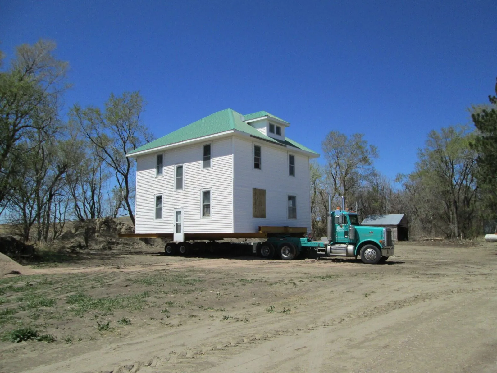 White farmhouse with a green roof on a trailer behind a teal truck, clear blue sky