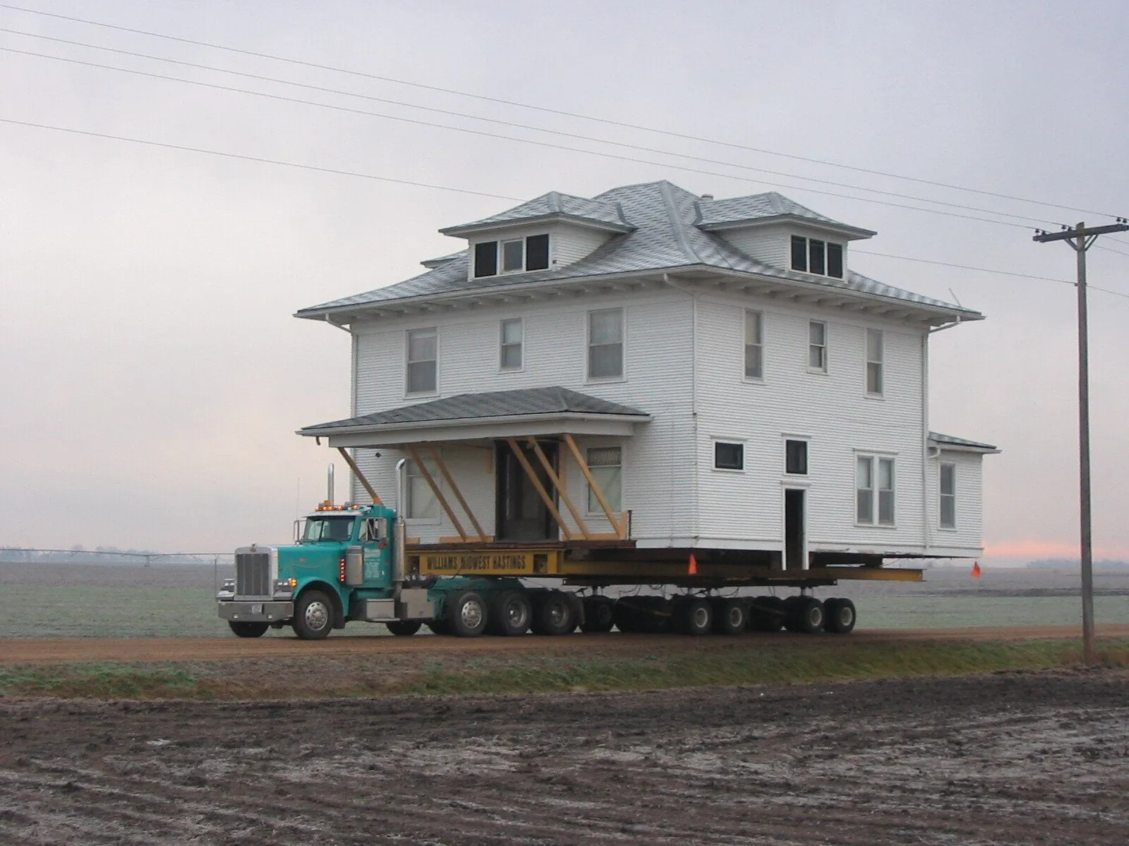 White two-story foursquare on a trailer behind a teal truck on the open prairie